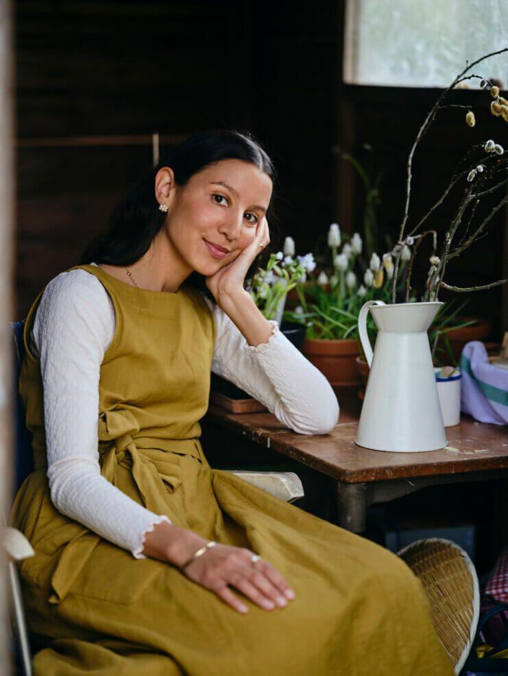 Johanna Tagada, sitting at a table with her head leaning in one hand, wearing a beige dress with a white top underneath