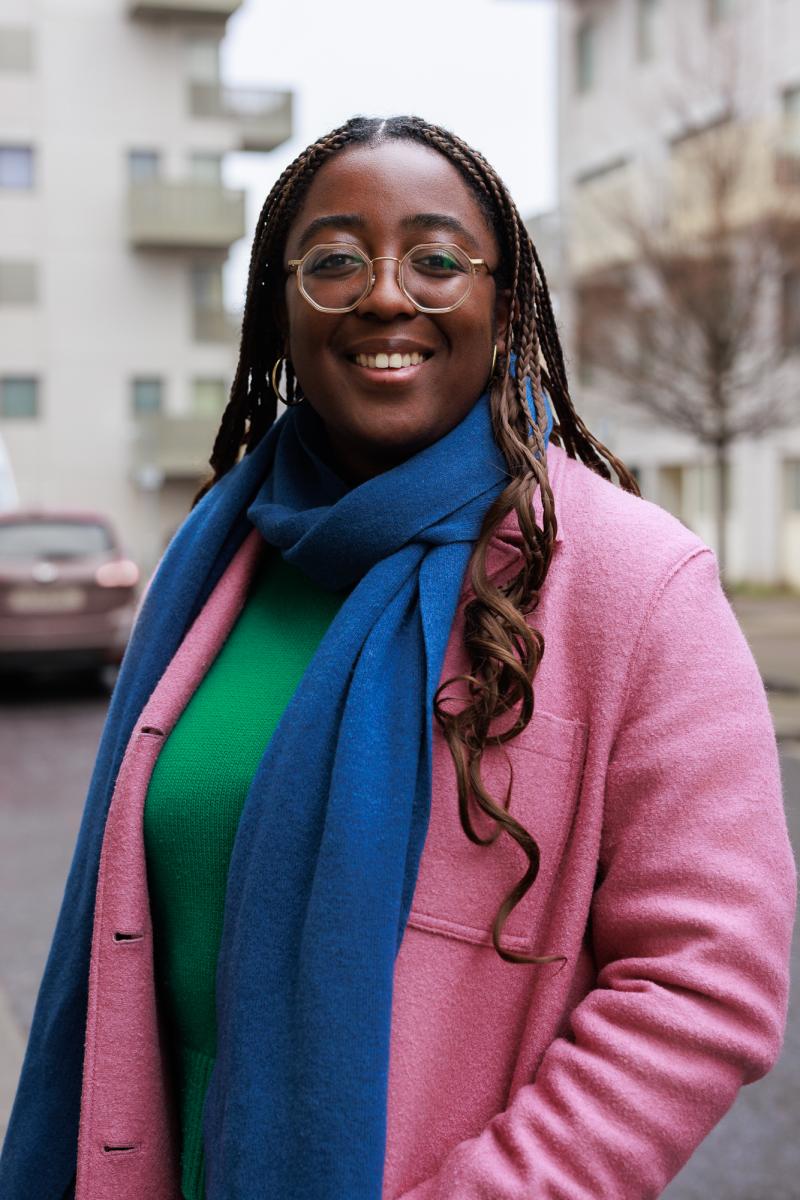 Betty Owoo smiling at the camera, wearing glasses, a blue scarf, green jumper and pink coat