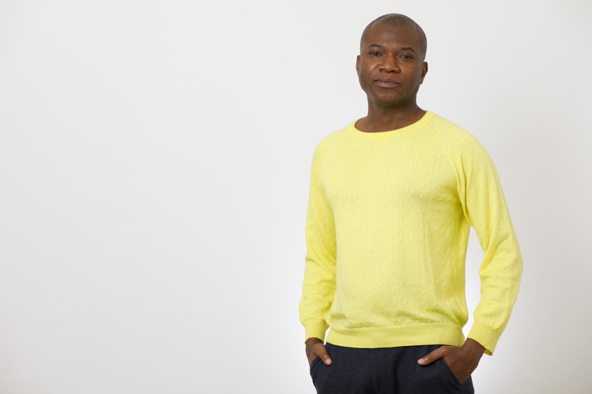 David Ogunmuyiwa stands facing the camera with his hands in his pockets, wearing a long sleeve yellow tshirt and navy trousers