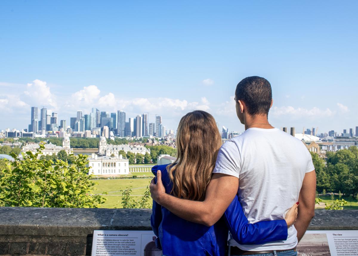 A man and a woman hug as they look out over historic Greenwich towards the skyscrapers of Canary Wharf. They have their backs to the camera