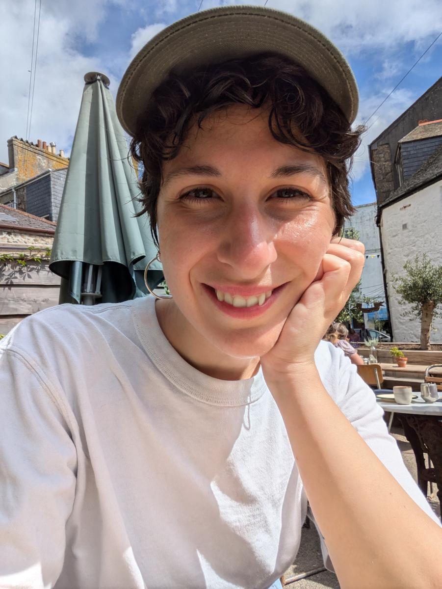 Ema Sala smiling at the camera with their head resting on their hand, wearing a white tshirt and beige cap