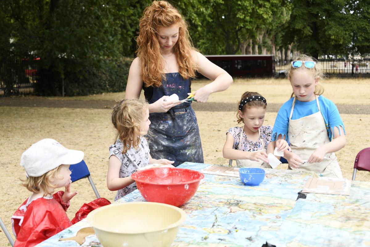 A group of children and an adult supervisor take part in a craft activity, with a table covered in a map of the world and bowls for children to put their hands into