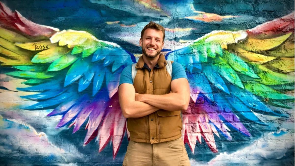 Sacha Coward stands with his hands crossed, wearing a brown gilet and blue shirt. He stands in the centre of some rainbow coloured wings painted on the wall