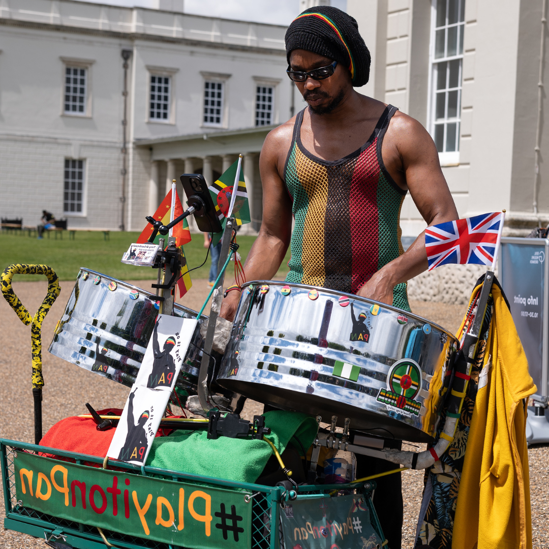 Man playing steel pans outside the Queen's House wearing a string vest with green, yellow, black, red and green vertical stripes