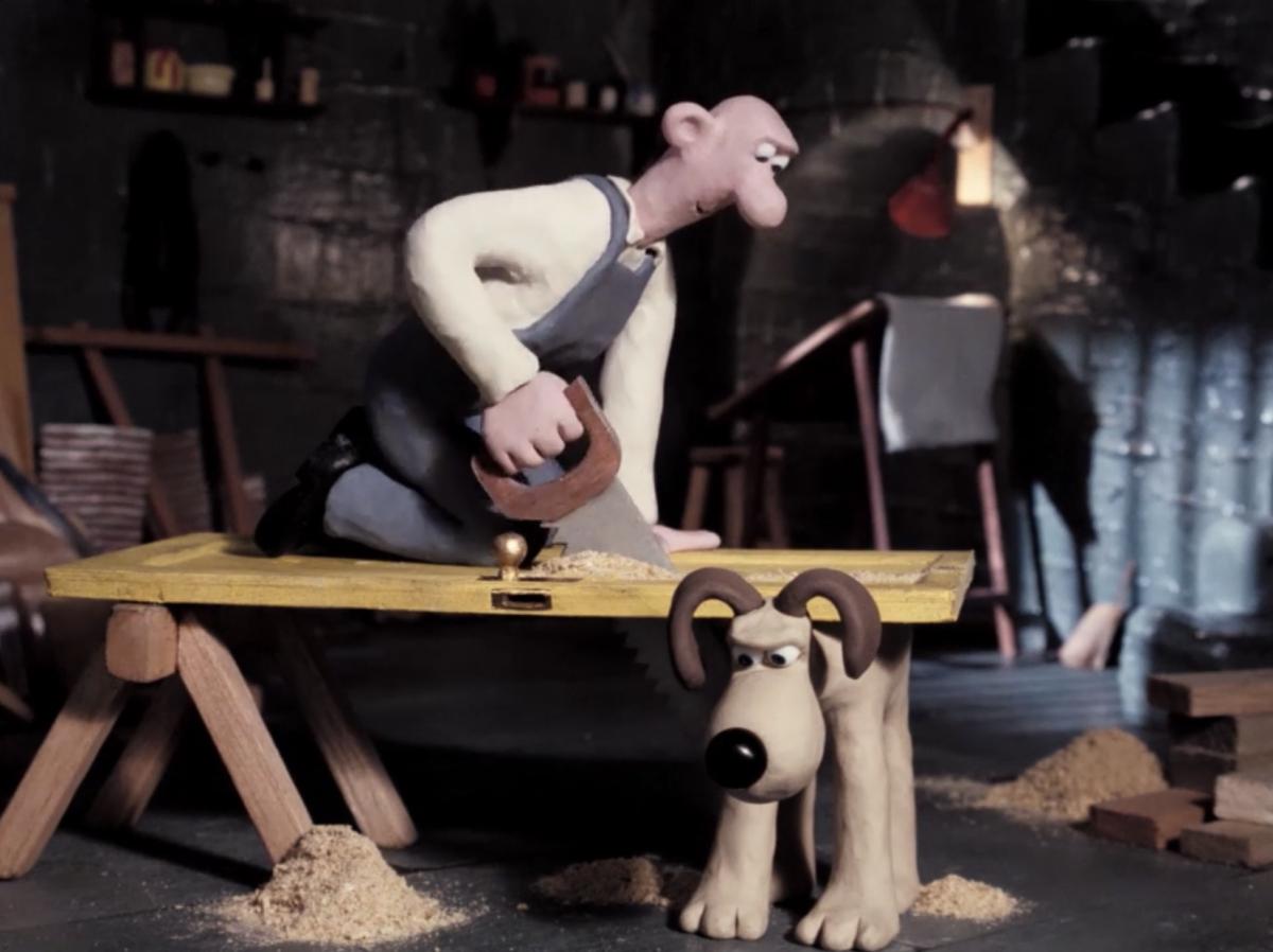 Still from Wallace and Gromit film A Grand Day Out where Wallace, a clay man, is kneeling on a door and sawing it propped up on a bench and also by Gromit, his dog