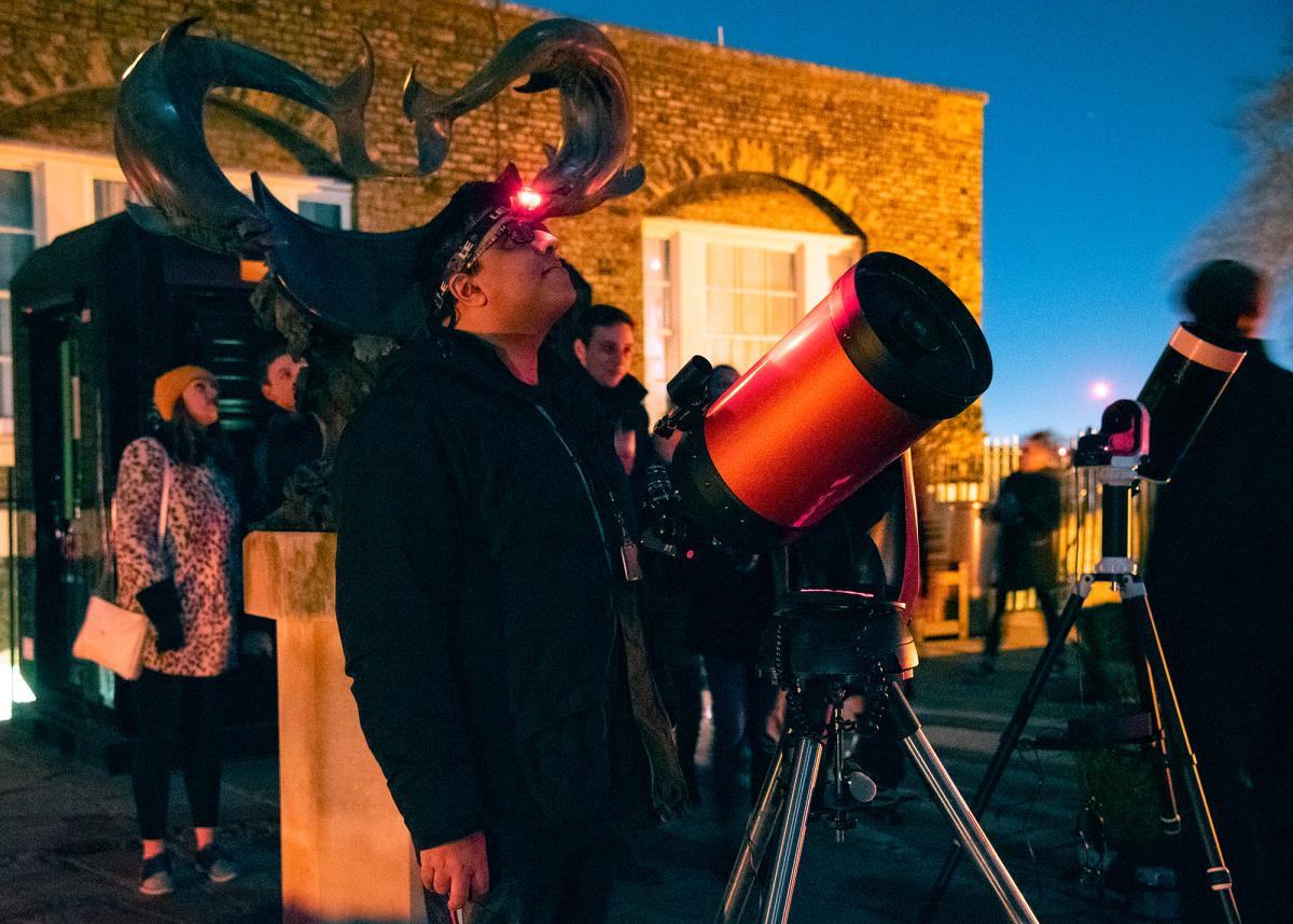 An evening astronomy session at the Royal Observatory, featuring a man stood next to a telescope and a group gathered behind