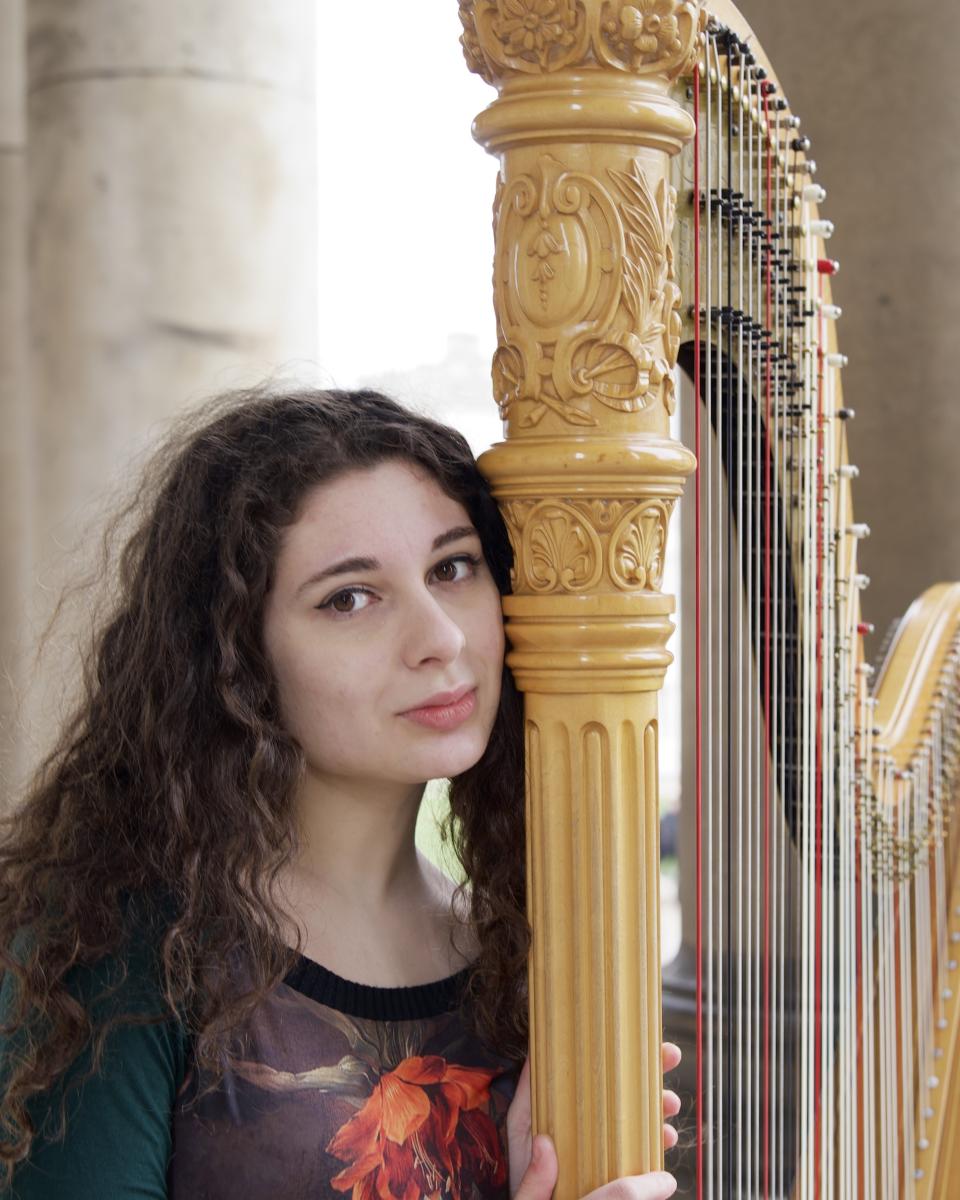 Profile photograph of harpist Lucia Foti. She has dark curly hair and her head is leaning against a large golden harp