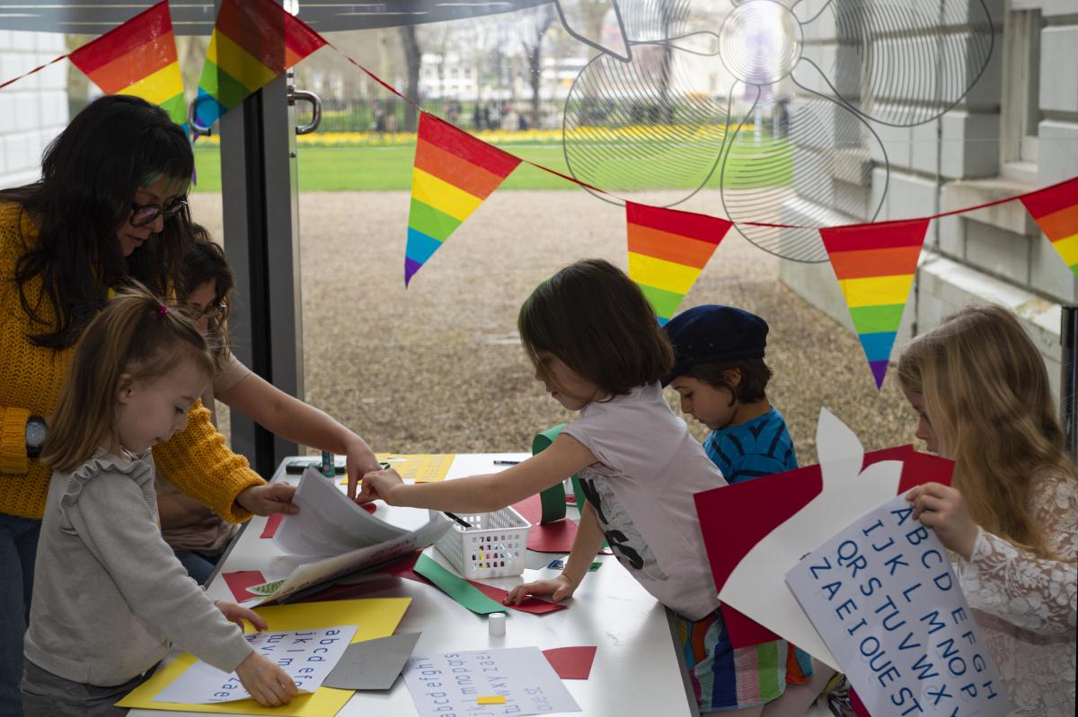 A group of children take part in a craft activity at a table covered in paper and letters of the alphabet. Bunting showing the Pride flag is in the background