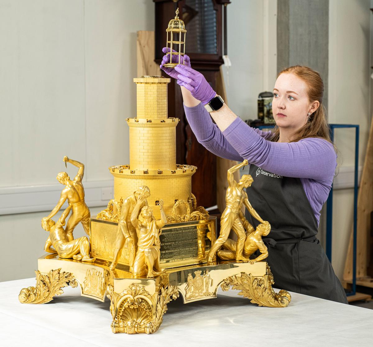 A museum conservator working on a large golden table centrepiece. The sculpture resembles a fortress, with three layers of ramparts surrounded by human figures and scenes of battle