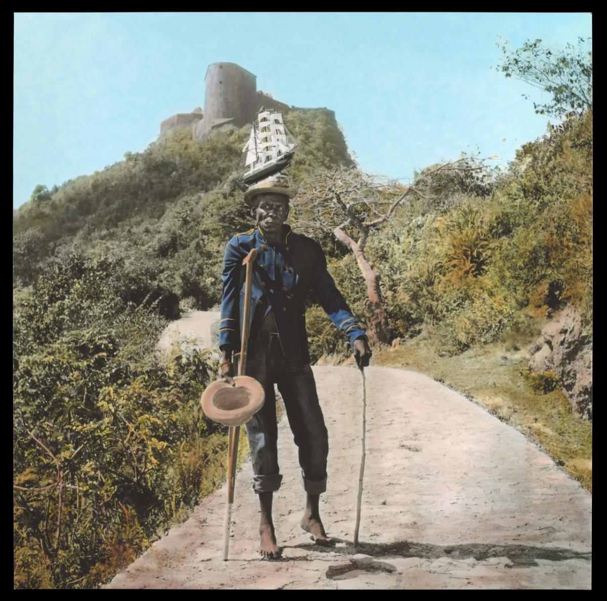 A man holding a crutch and wearing a hat stands on a rocky road by a hillside