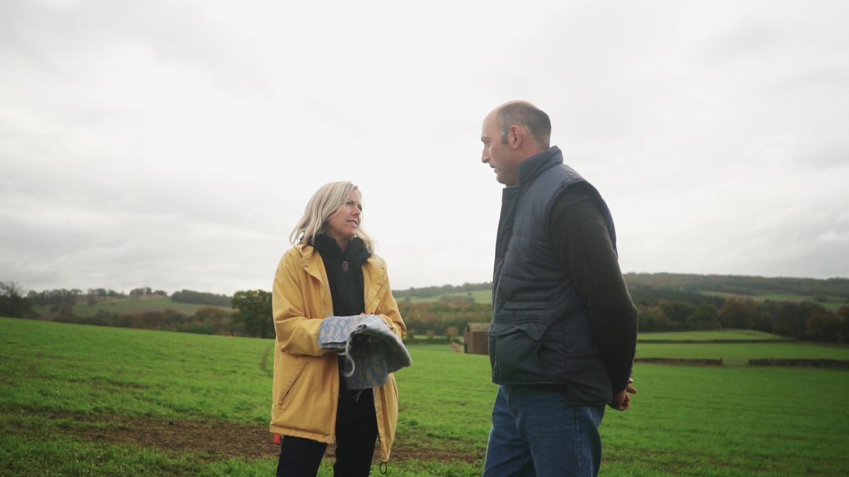 A still from the film Fabric of the Land: The Untold Yarn, showing artist Ella Doran examining a piece of textile design with a farmer in a field