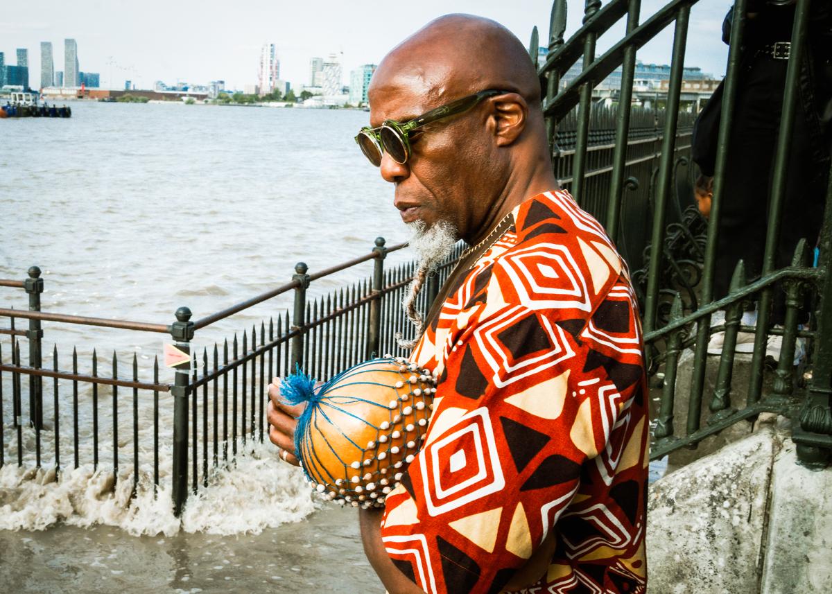 A Black man wearing green sunglasses and a geometric patterned top stands contemplatively by the River Thames