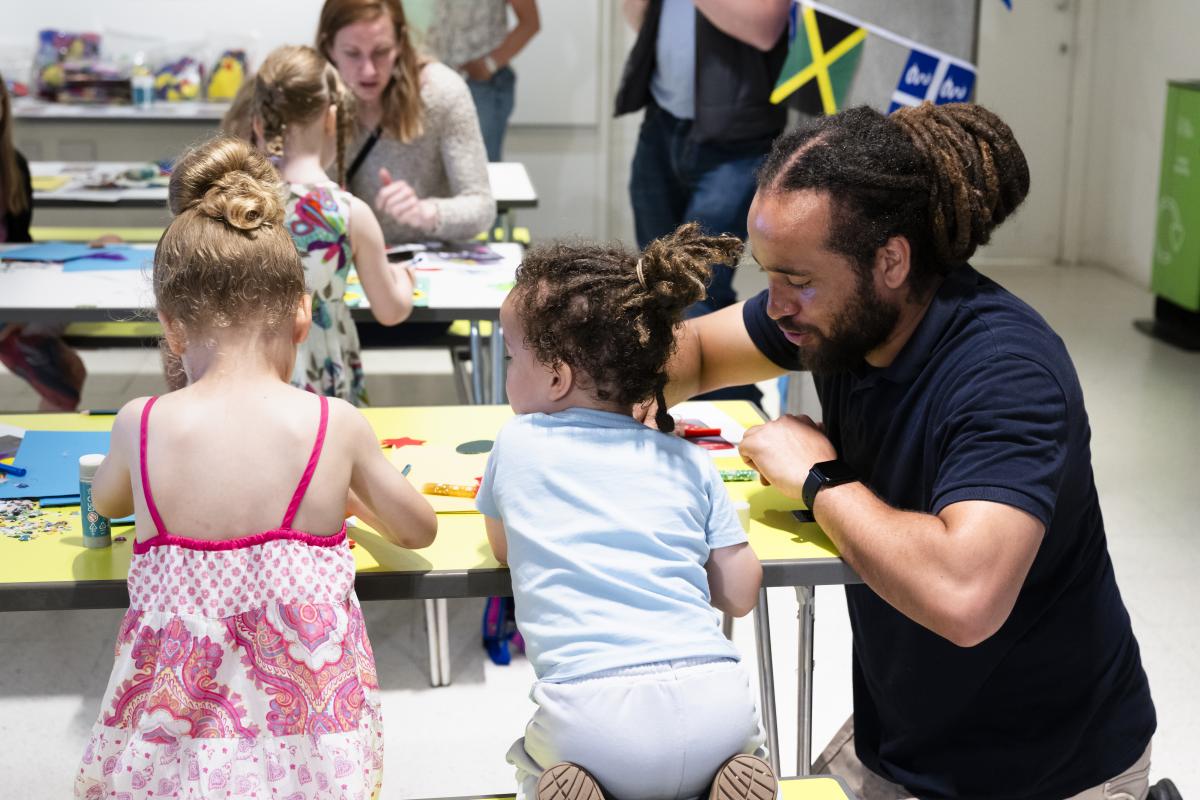 Photo of man crouching next to a table that two children are sitting at and making something