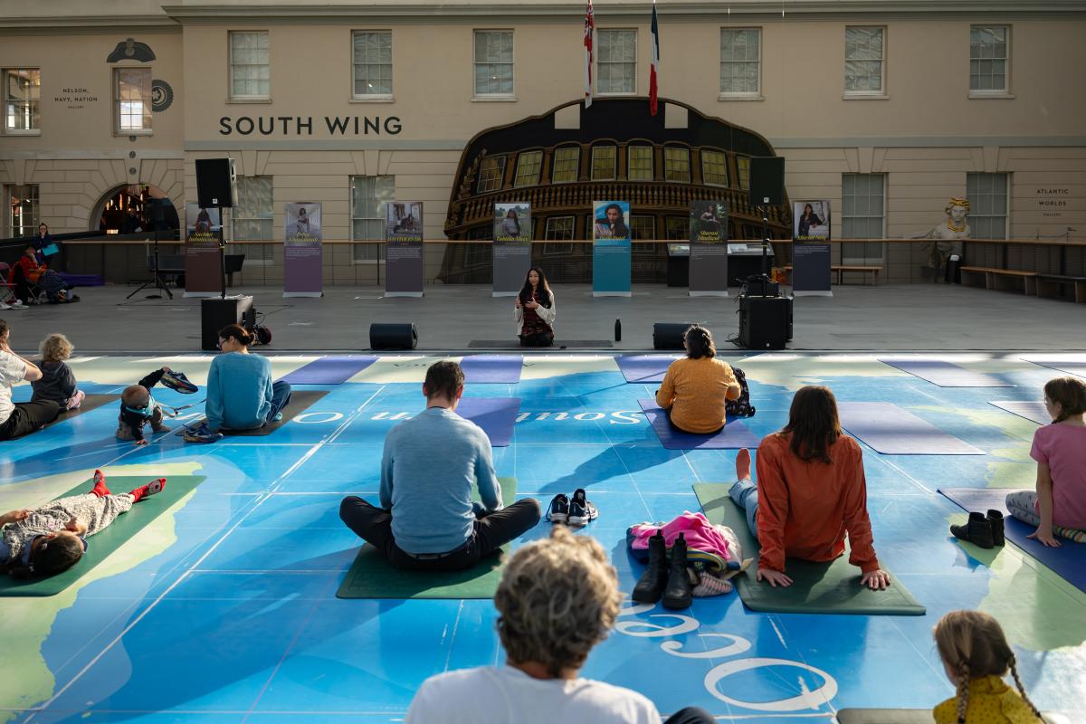 Group of people sitting facing away from camera on yoga mats on the floor which is a giant map, looking towards instructor