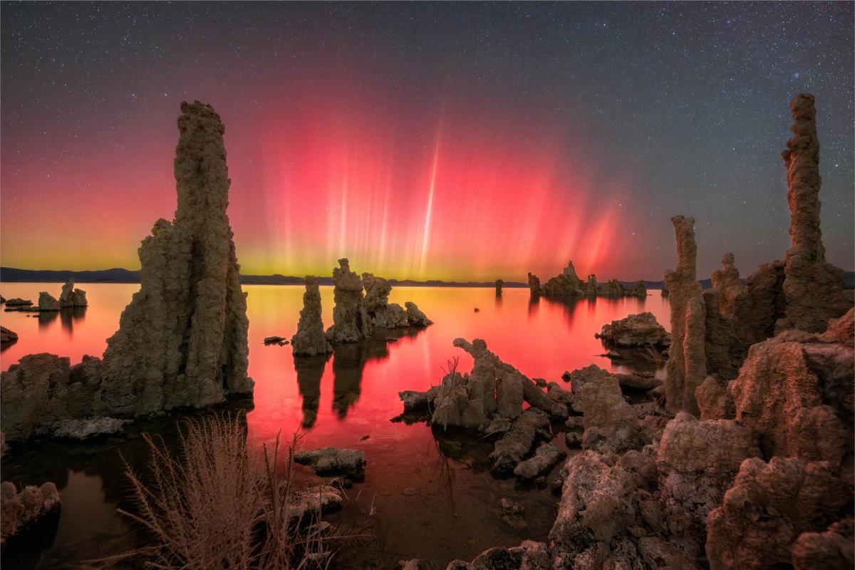 Photo showing landscape, on the bottom half is a lake with rock protrusions and in the top half is the sky with bright green and purple aurorae, which is reflected in the lake