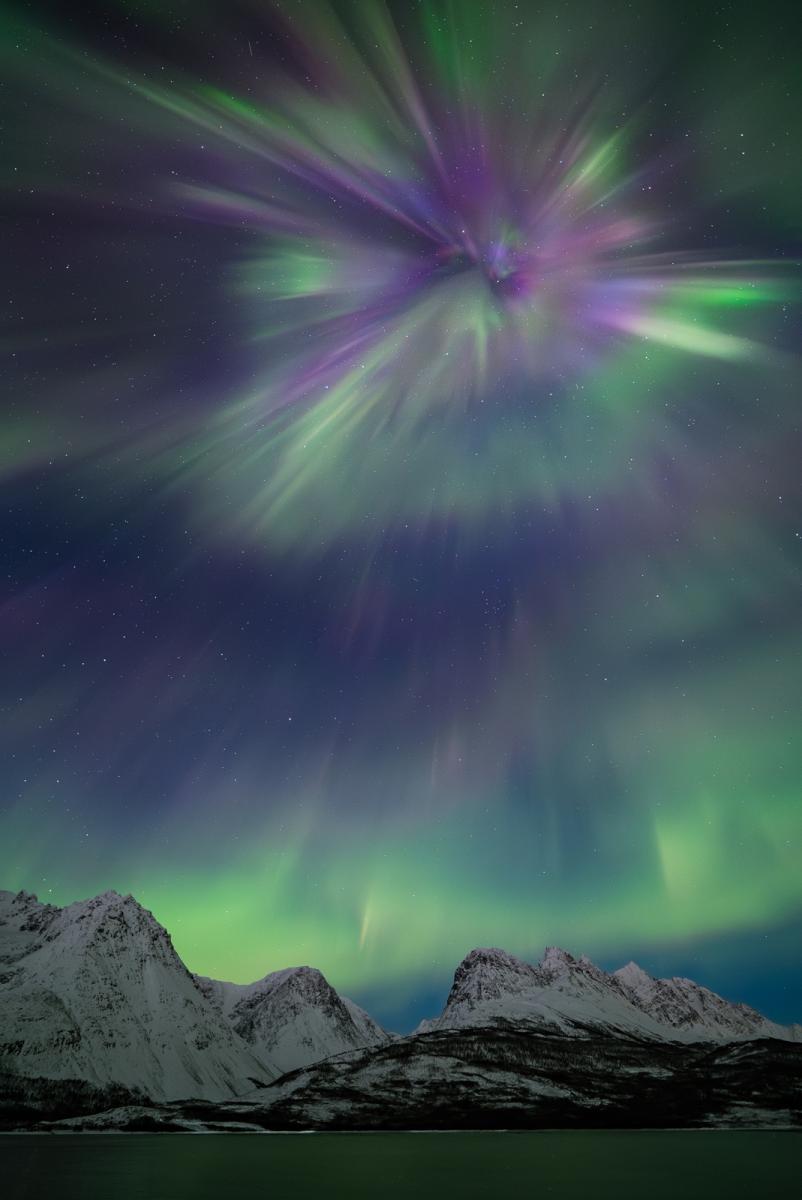 Photo showing icy snowy mountain landscape with vast sky above filled with green and purple aurorae, at the top is a large firework-shape aurora in green and purple