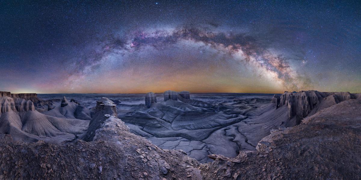 Desolate, dry and cracked Utah landscape, below a starry Milky Way arching in the sky in purples, blues and oranges