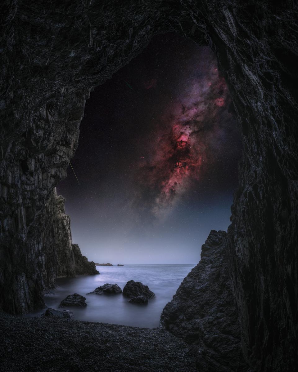 Photo taken from inside a cave looking out on a seascape with the Milky Way diagonal in the sky above