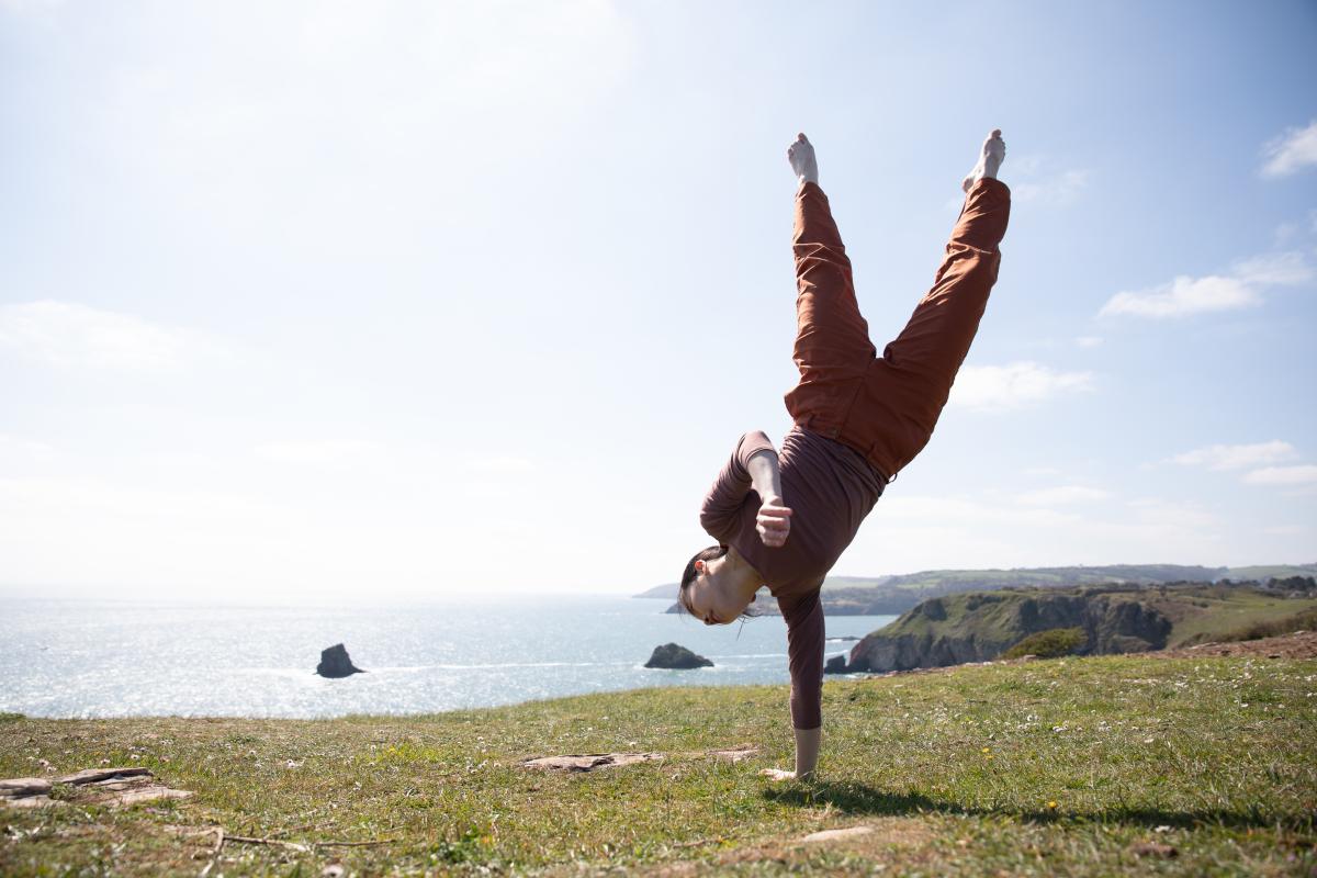 A dancer does a one-handed handstand with a seascape behind