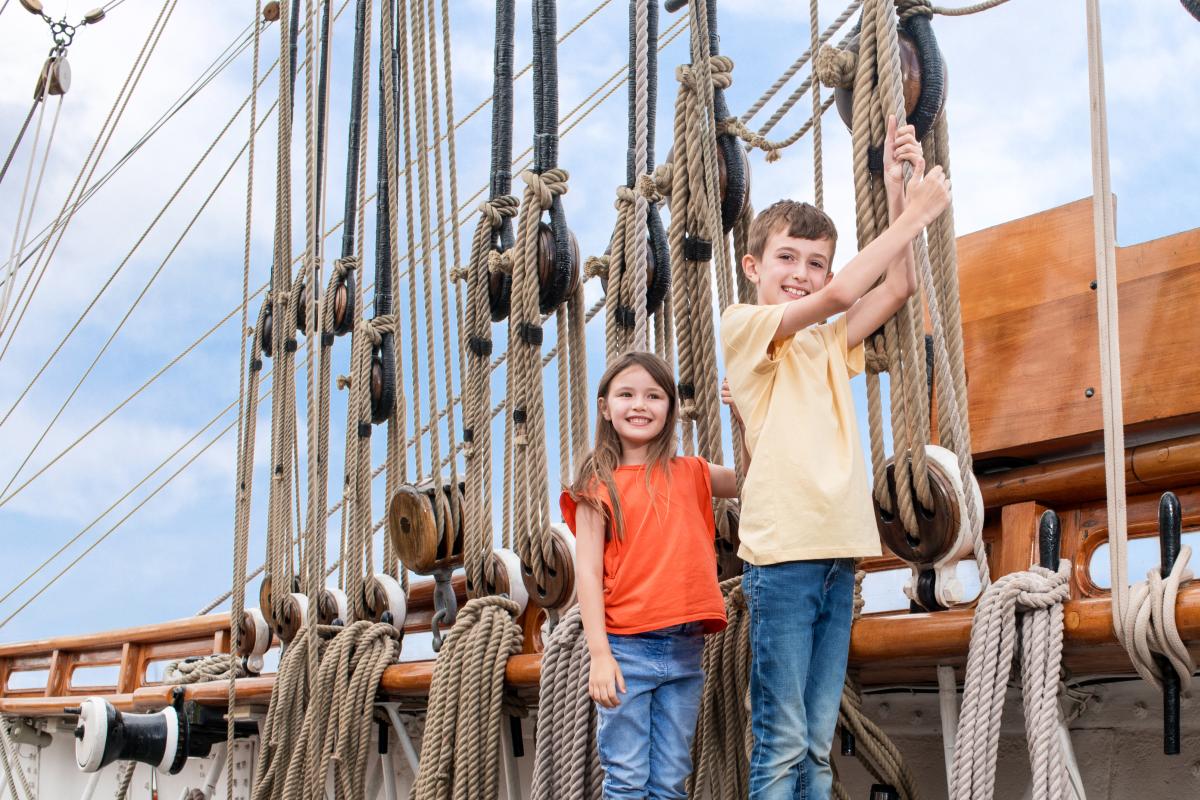 Photo of kids on a historic ship, a younger girl and older boy holding onto ropes that make up the rigging of the ship and smiling
