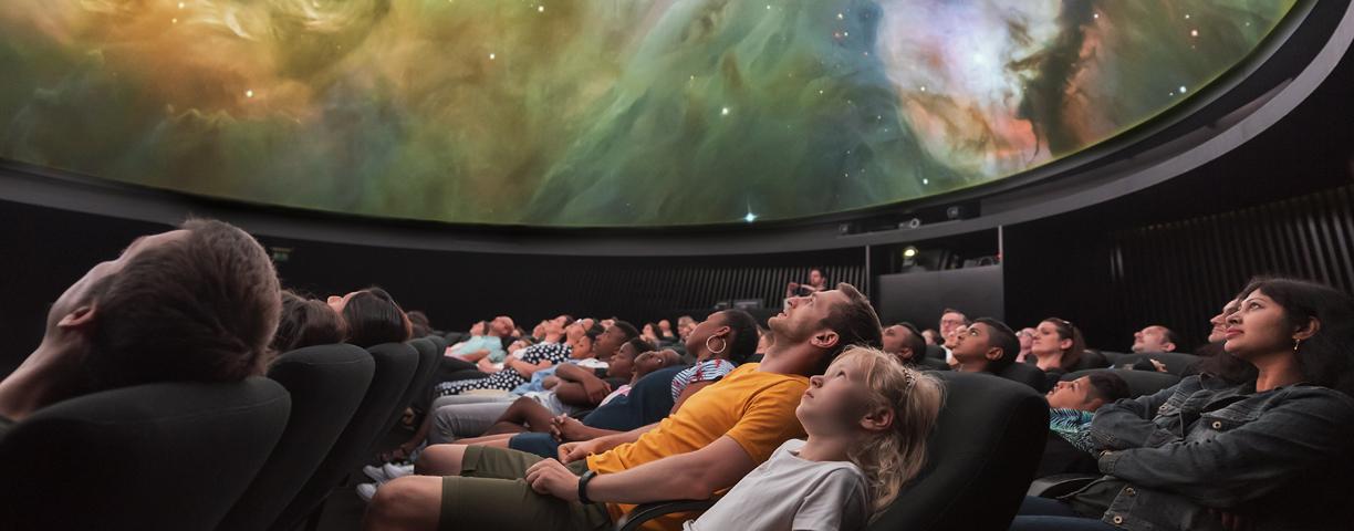 A large group of people, watching a planetarium show. The screen is showing a swirl of a galaxy