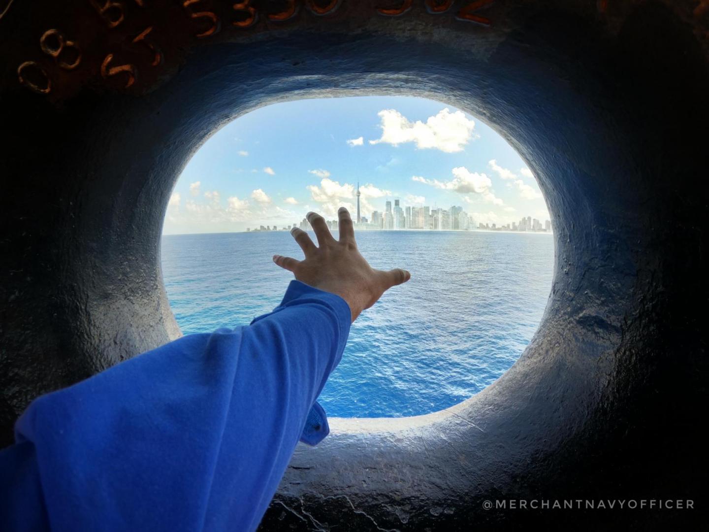A sailor on board holds his hand out the porthole to reach for the shore and buildings