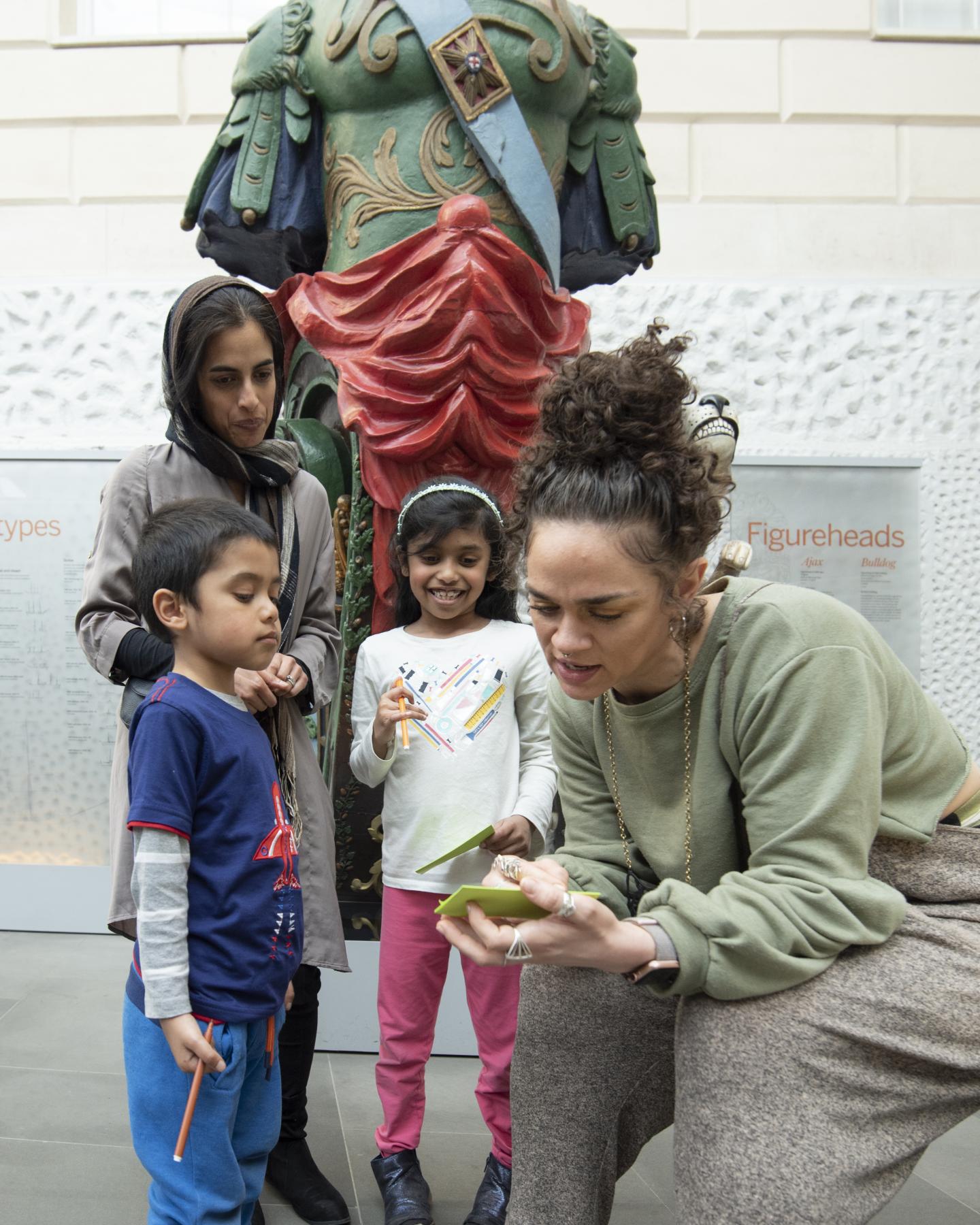 A group of adults and children are reading from a piece of paper in front of a figurehead. 