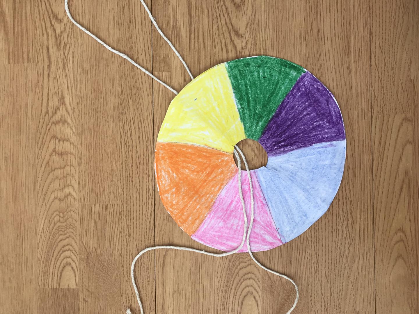 a doughnut shaped piece of card with 6 sections coloured in using blue, pink, orange, yellow, green and purple. A loop of string has been pushed through the hole in the centre.  