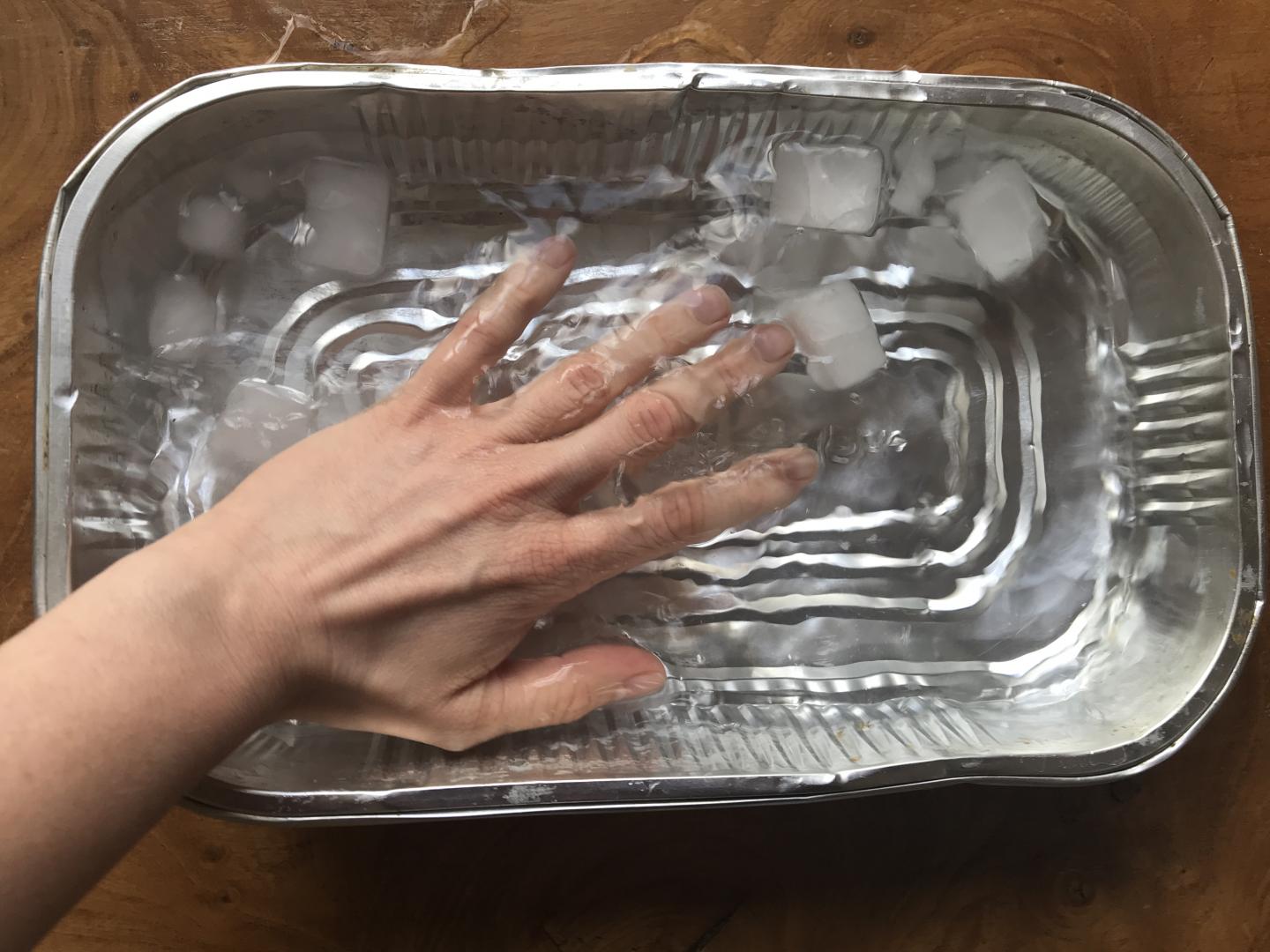 Water and ice cubes are in a shallow dish. Someone's hand in making waves.