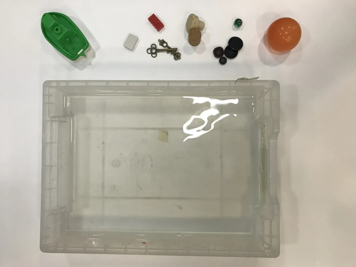 a tray of water with different objects including a toy boat, keys and corks nearby.