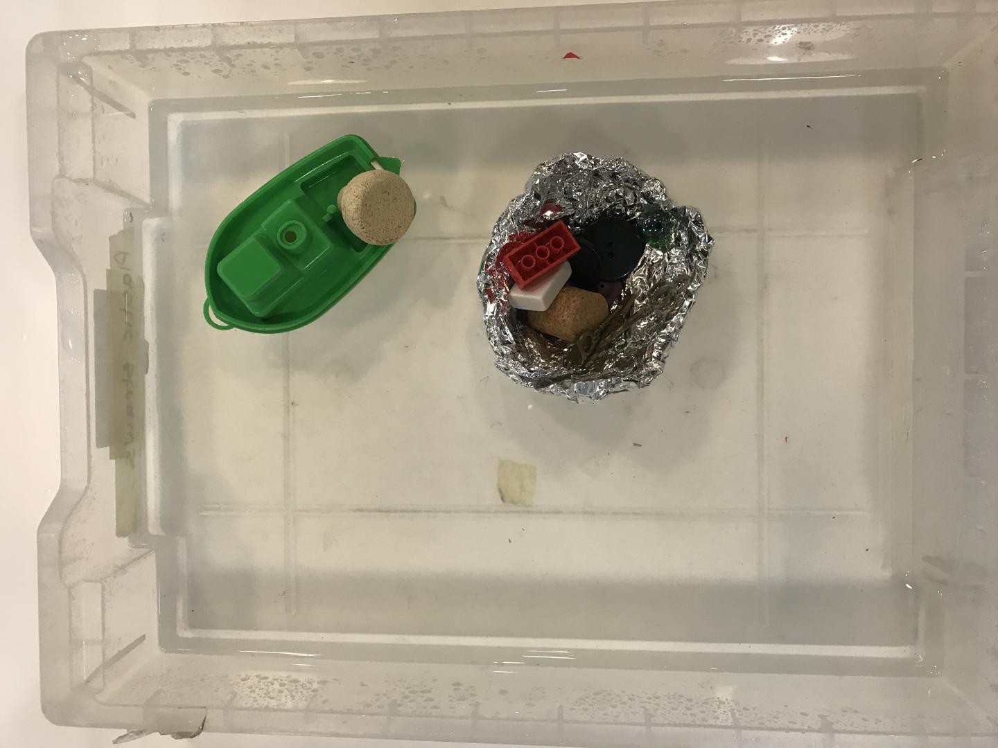 A toy boat is floating in a tray of water with a cork on the back. Another boat has been made from tin fil and has things like lego and buttons inside it. 