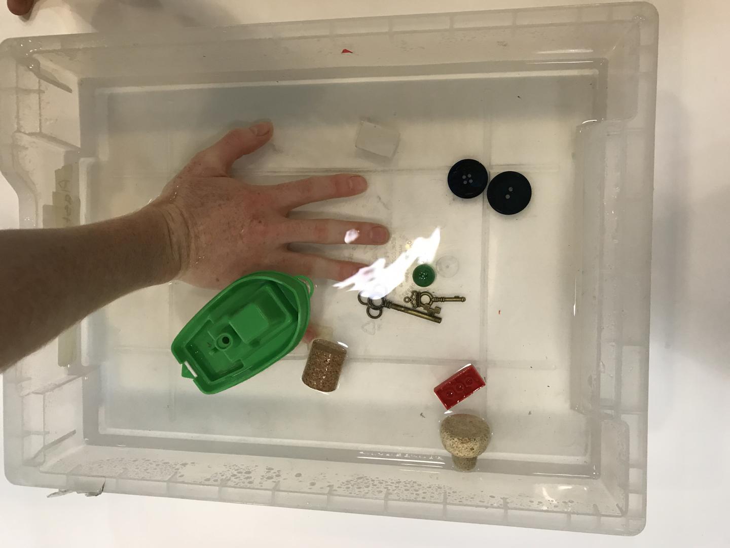 Someone is plunging their hand into a tray of water. Also in the water are a toy boat, some corks and keys, and other miscellaneous objects. 