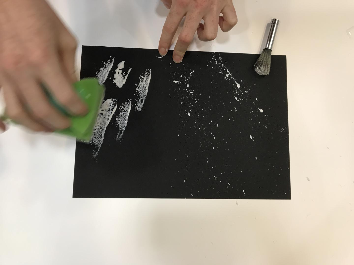 White paint has been flicked onto black card and someone is using a sponge to add more white paint.