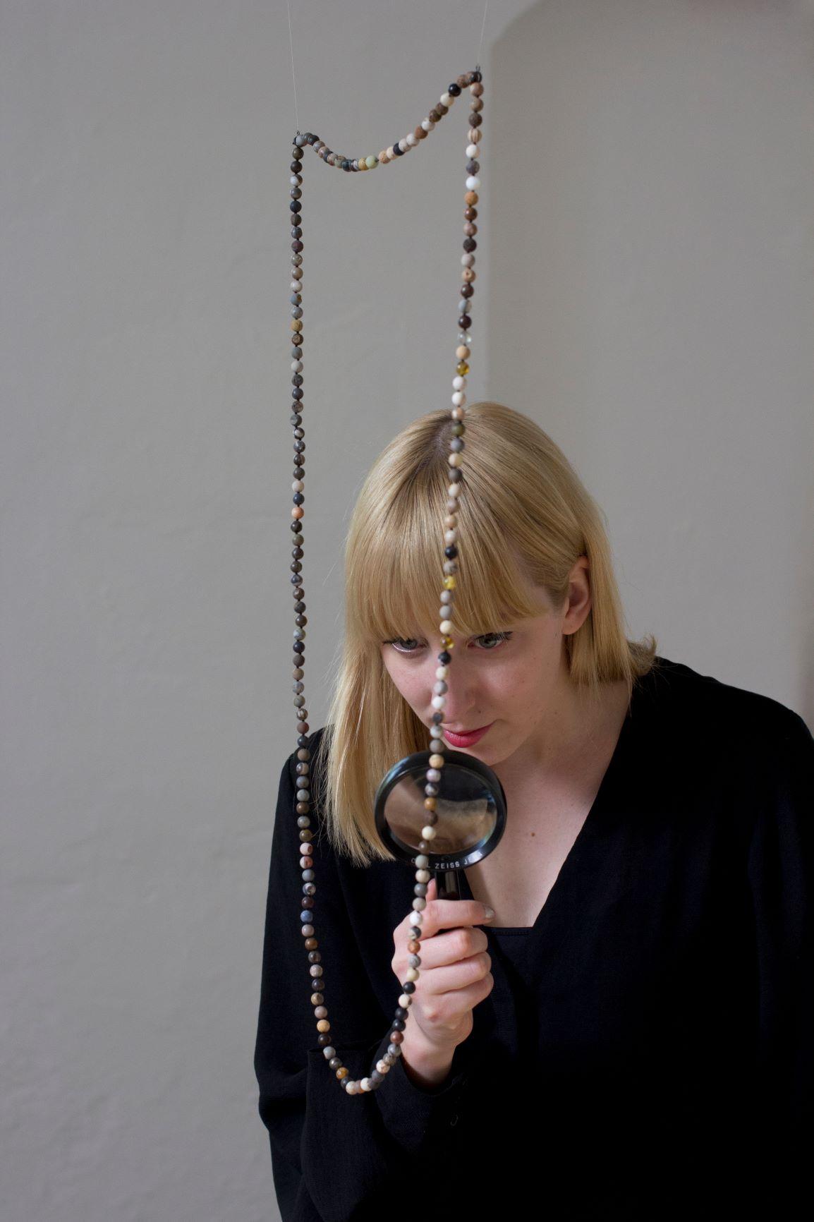 Artist Katie Paterson looks through a magnifying glass at her work Fossil Necklace, a necklace comprising 170 fossils carved into spherical beads