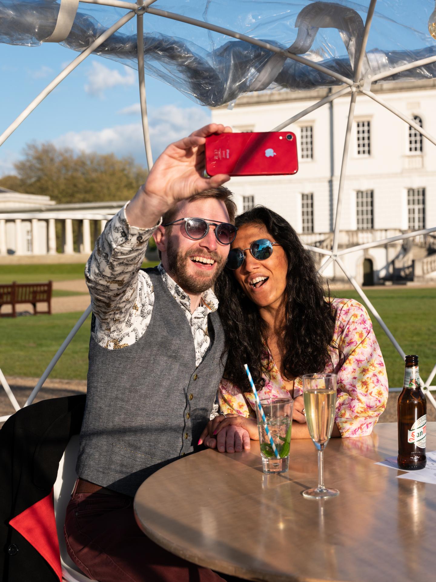 A couple takes a selfie in the Queen's House Dining Domes, with drinks on the table and the Queen's House in the background