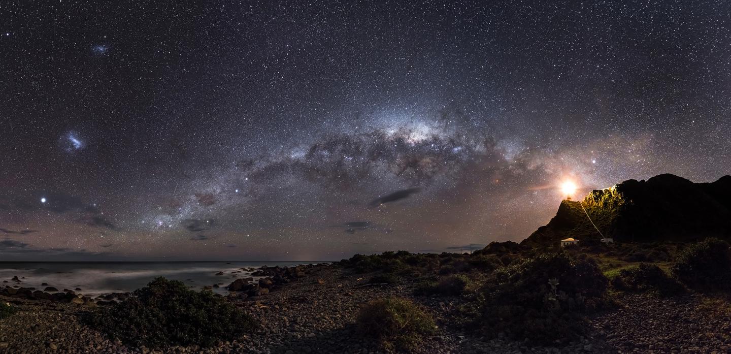 A skyscape at night showing a constellation of stars appearing to point towards a bright lighthouse on the right