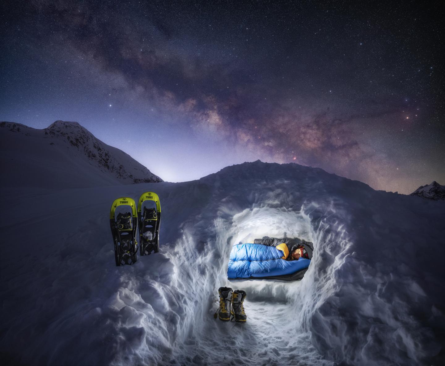 Man wrapped up in a sleeping bag in an igloo with shoes outside and the Milk Way in the sky