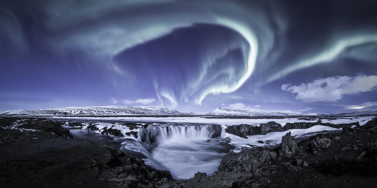 A landscape photo of a waterfall with the green curtain of an aurora dancing in the sky above