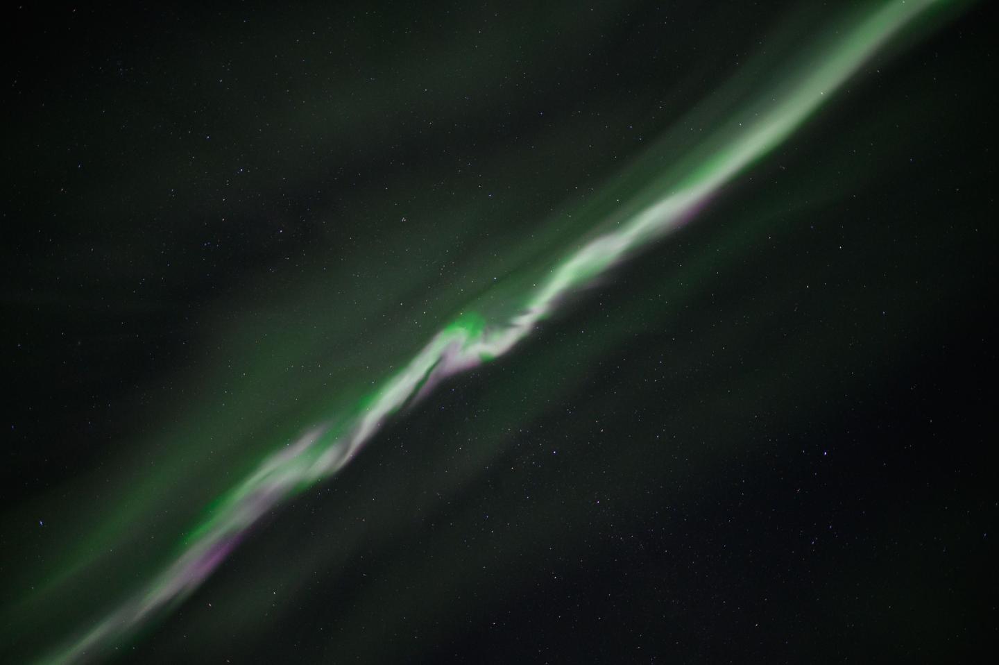 A thin blurred line of green and purple aurorae cuts diagonally across a dark sky