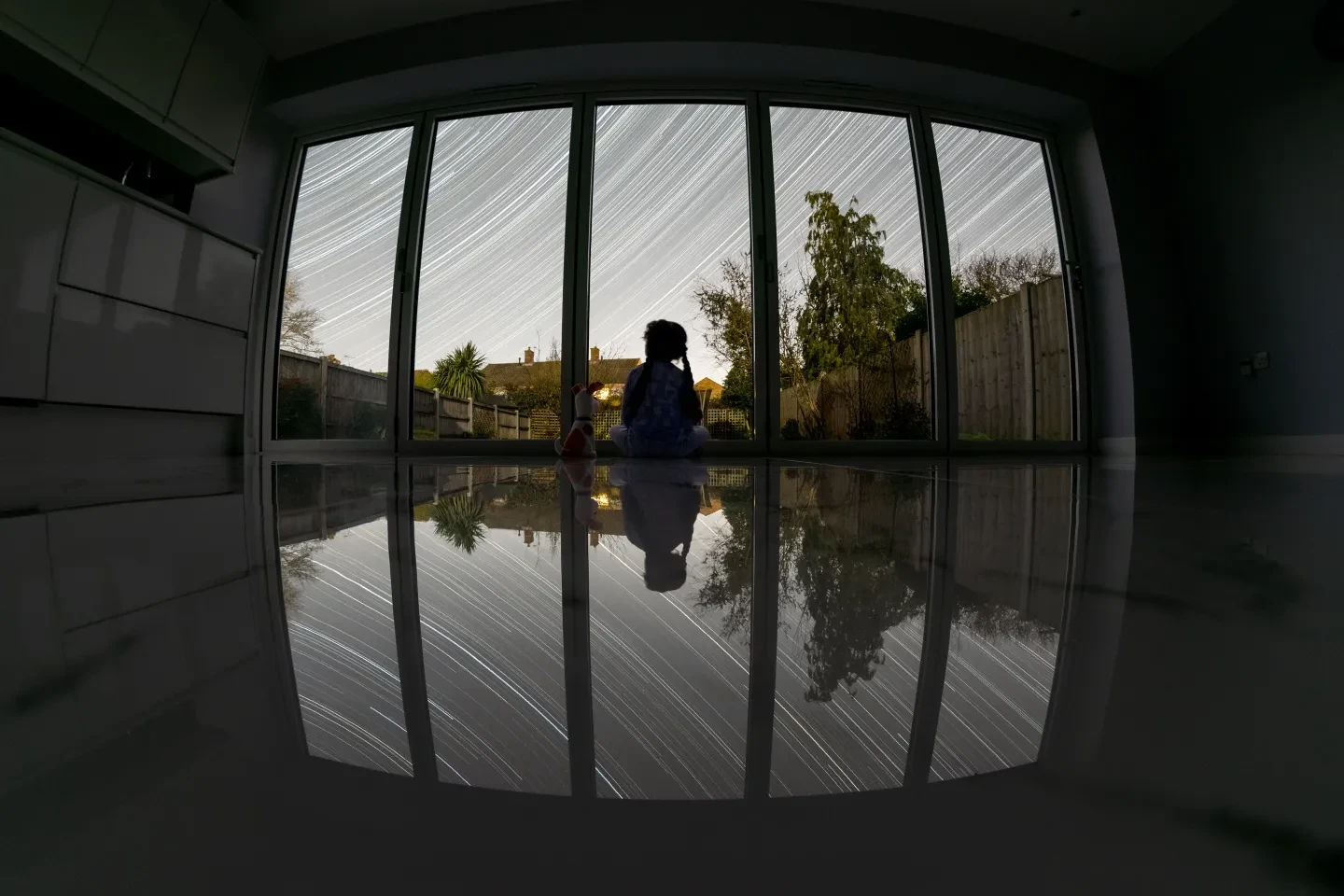 A girl sits behind bi-folding doors overlooking a garden. Star trails cross the sky above