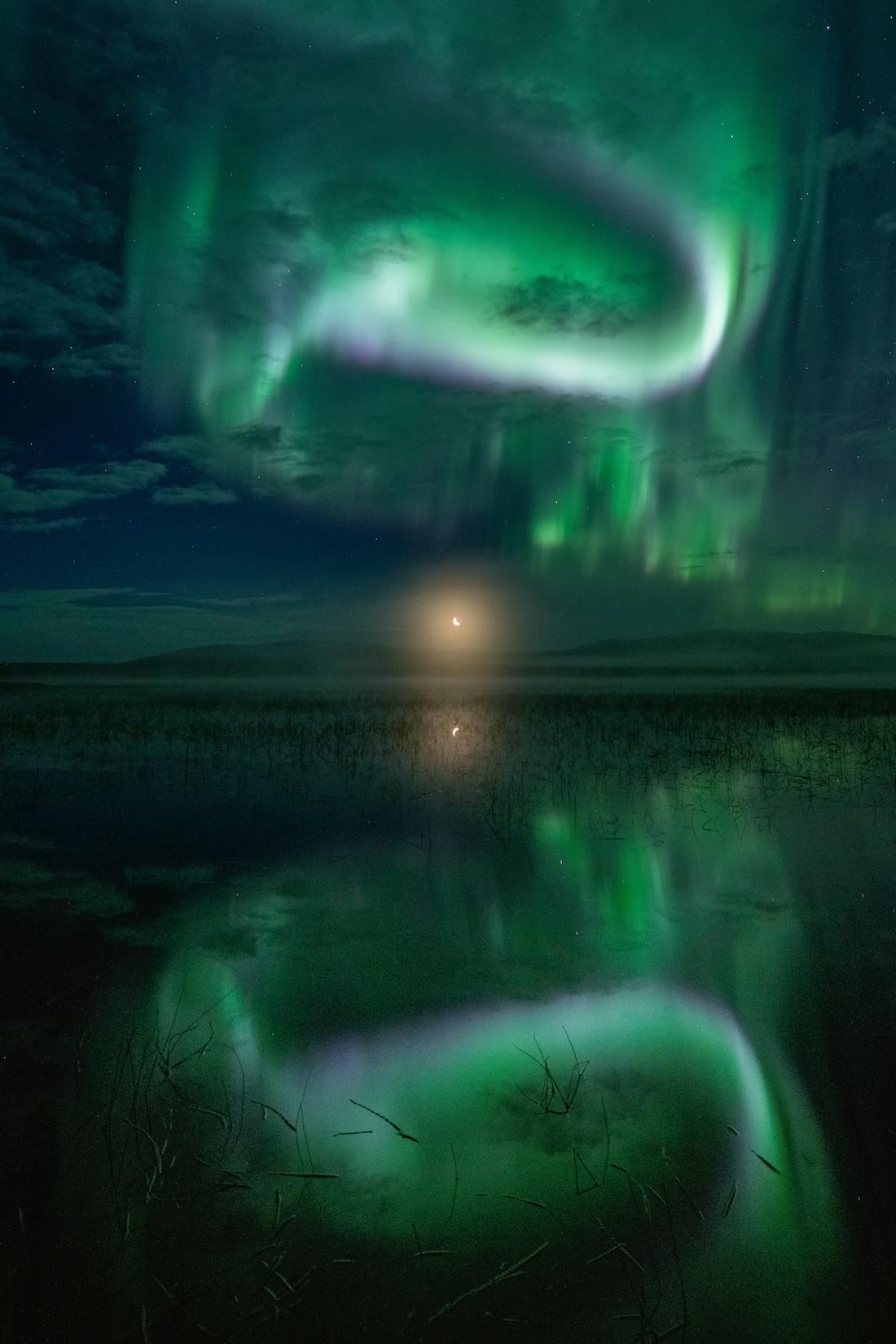 A green aurora reflected in water, with the Moon rising in the centre