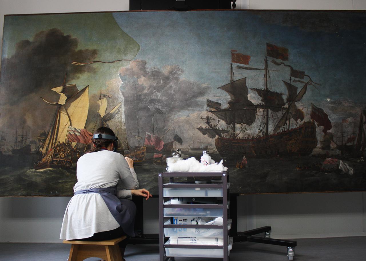 A paintings conservator works on a giant oil painting of a maritime scene