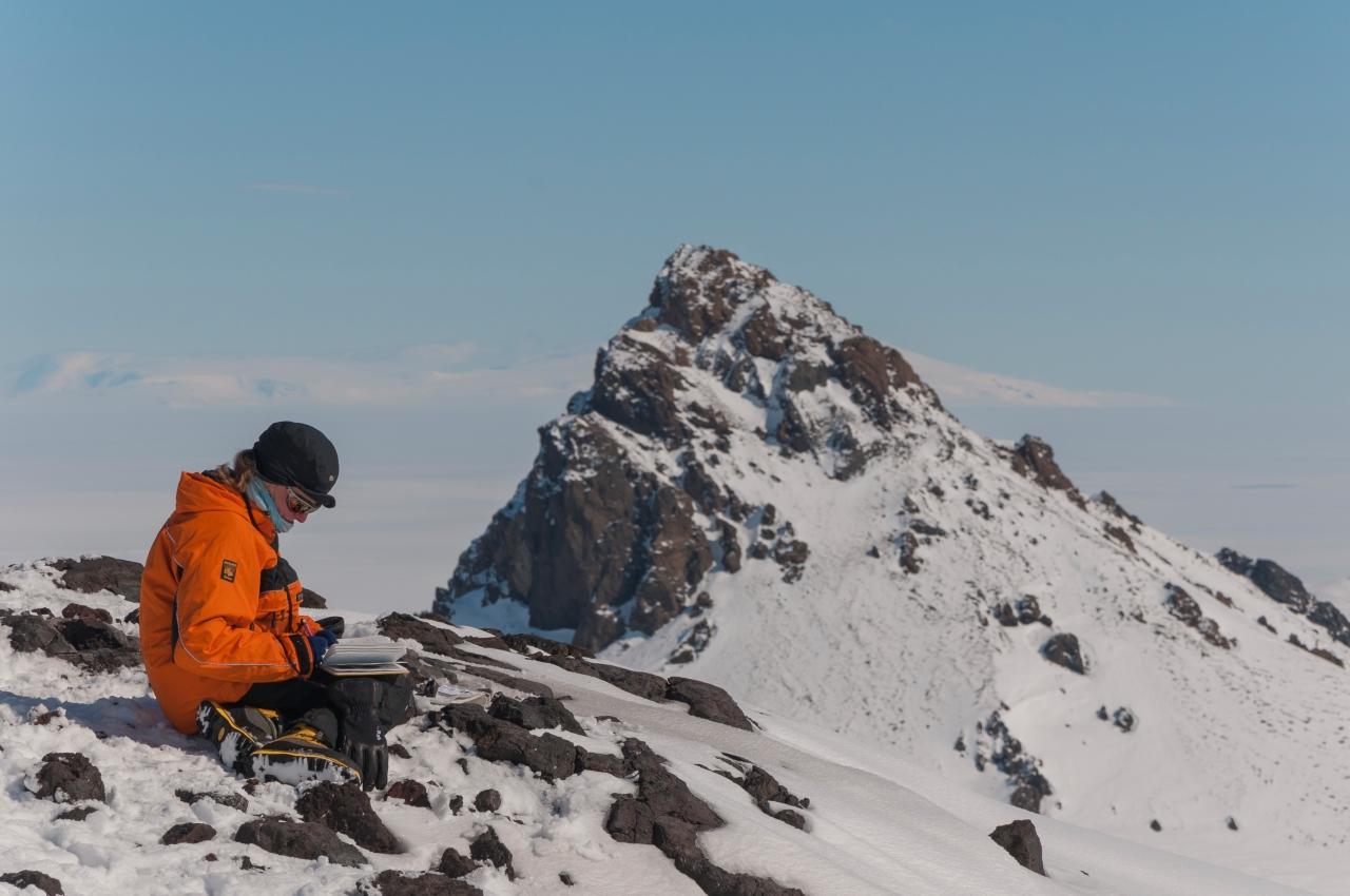 A scientist takes a note while on a research trip to Antarctica. She is sitting on a rocky outcrop with a snowy mountain in the background