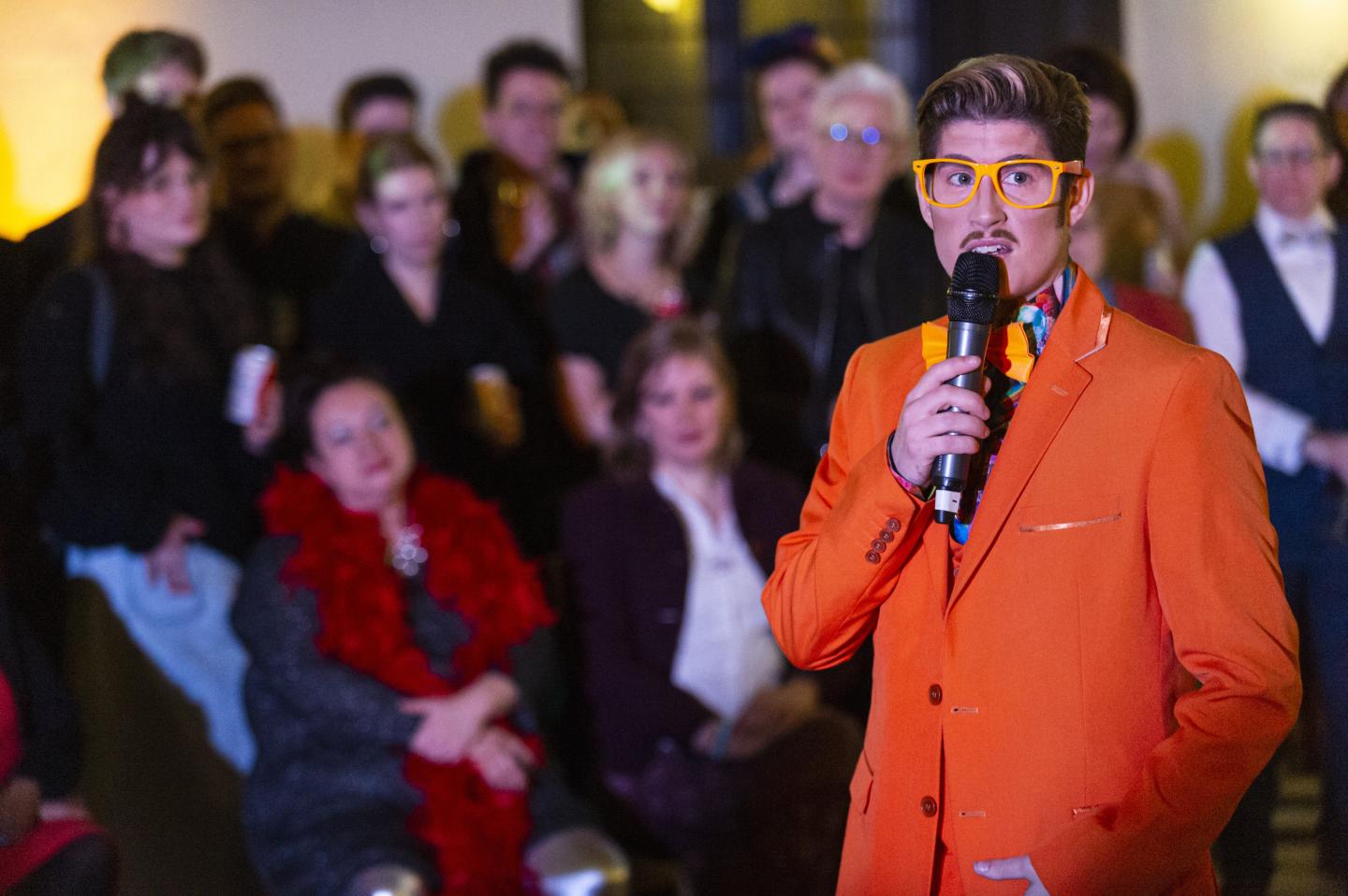 Adam All wears a bright orange suit with matching glasses and a bow tie and speaks into a microphone with an audience behind them