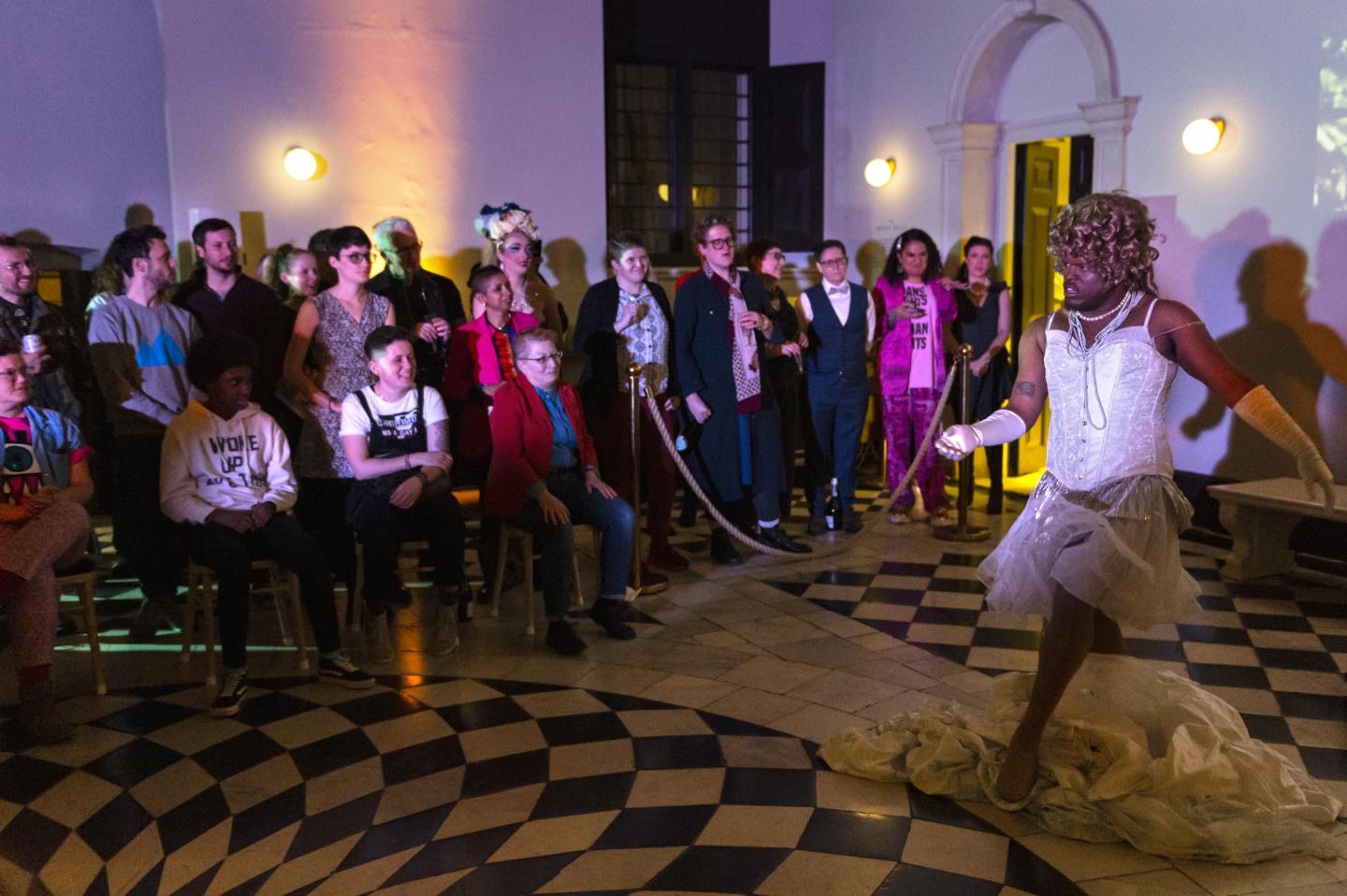 A photo of Lasana Shabazz in their Queen Charlotte outfit striding through the Great Hall in the Queen's House with an audience in the background. Taken at the Fierce Queens event in 2020.