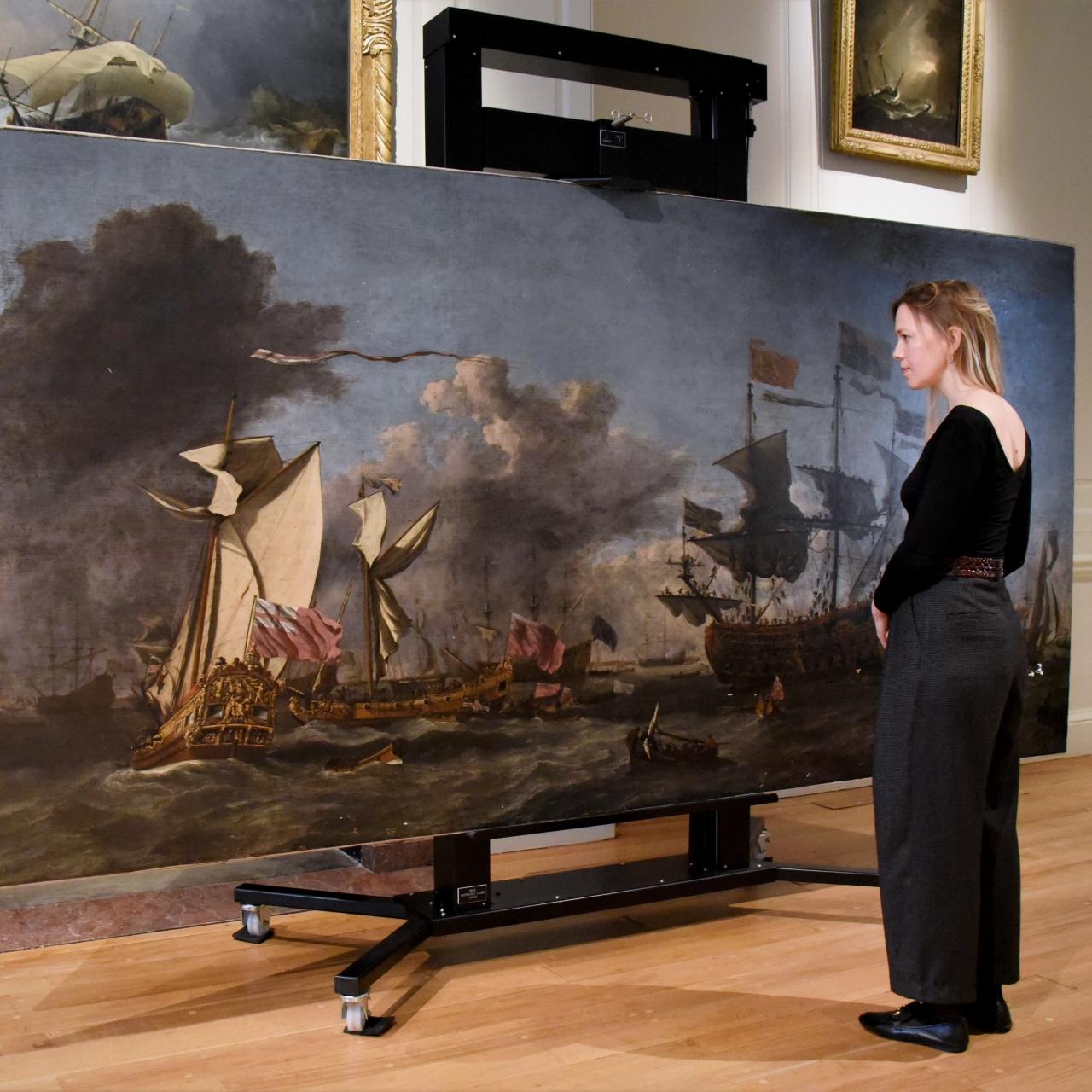 A woman looks at a large oil painting showing a naval scene. The painting is displayed on an easel