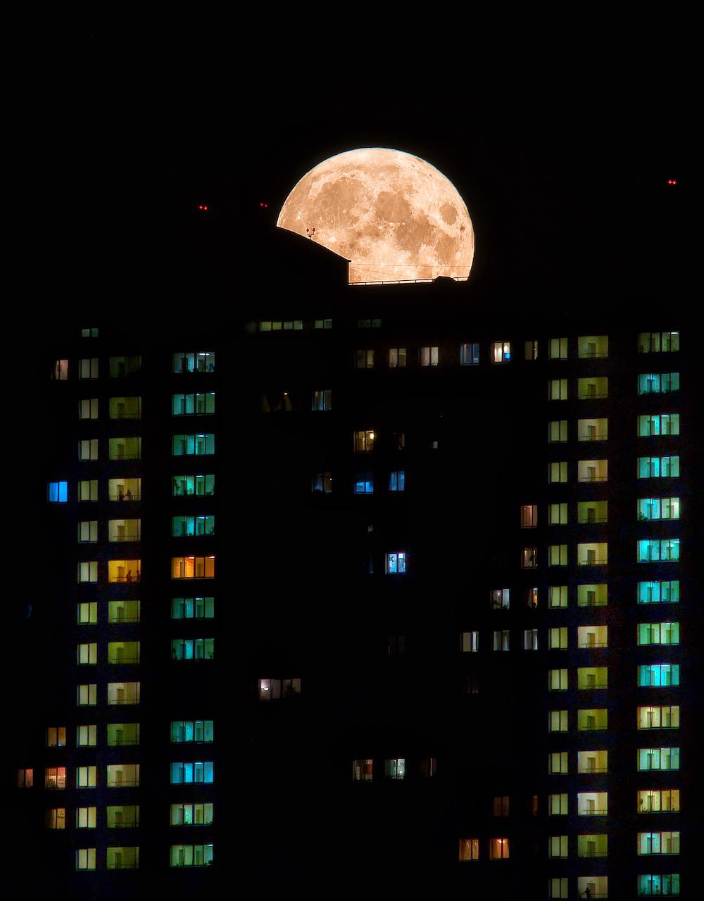 A full Moon appears over a block of flats lit up at night