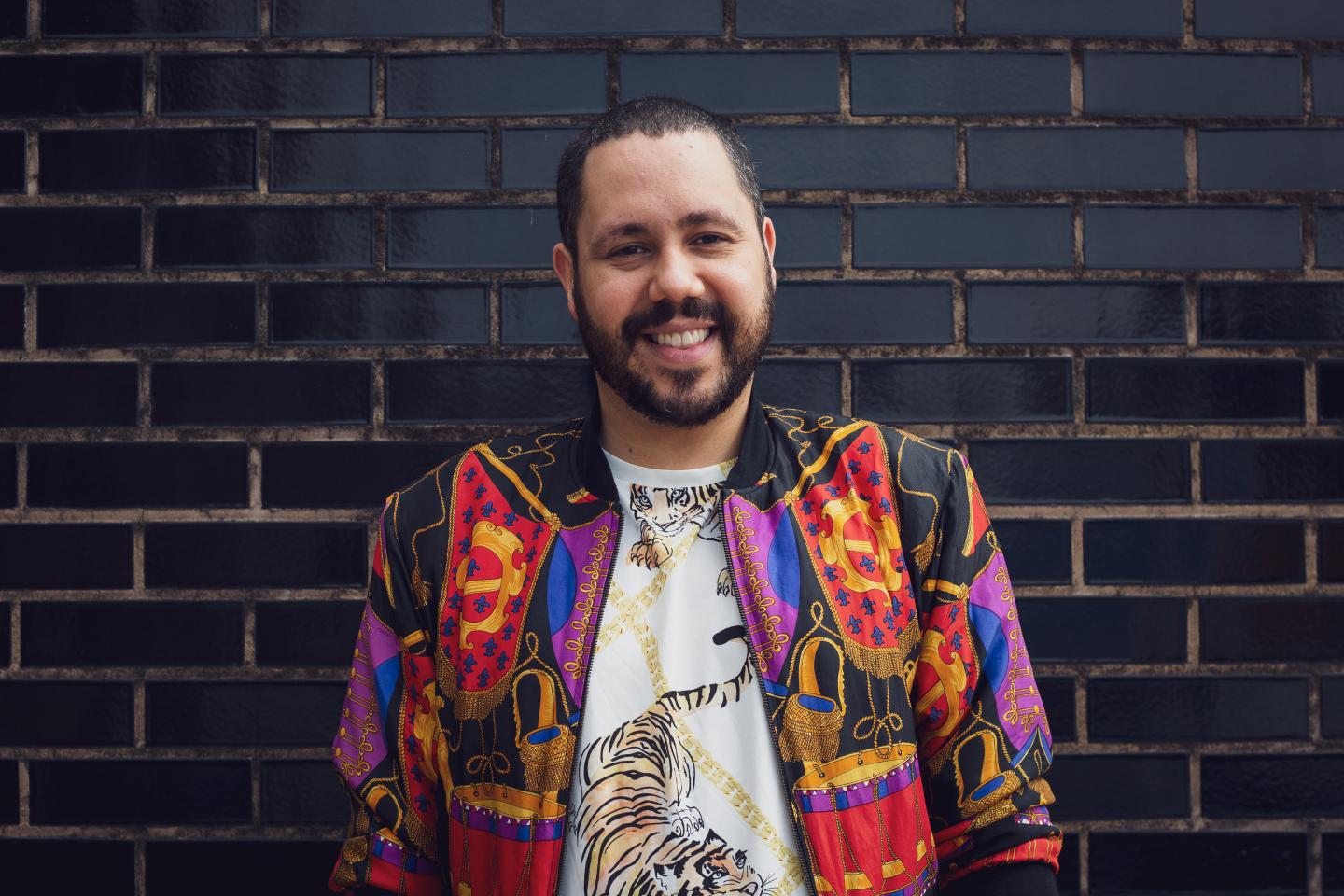 Adam stands in front of a black brick wall wearing a multi-coloured jacket and white t-shirt with a tiger on it
