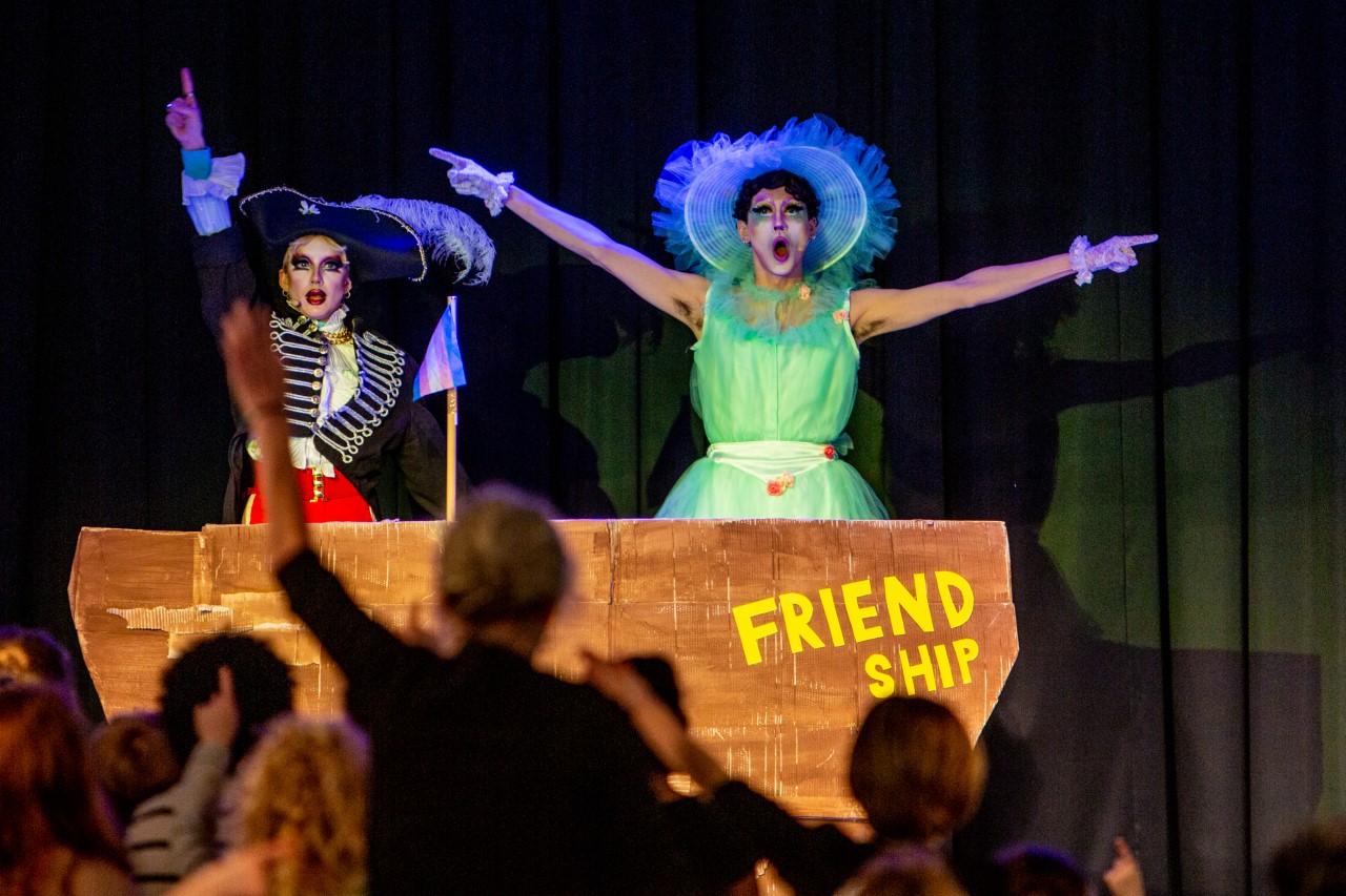 ShayShay and Sophie Brain stand inside a make-shift boat entitled 'Friendship'. ShayShay is wearing a frilly light green dress and white wide-brimmed hat with their arms in the air. Sophie Brain is wearing a ship captain's outfit and is pointing up. An audience of children stand in front of them with their arms in the air.