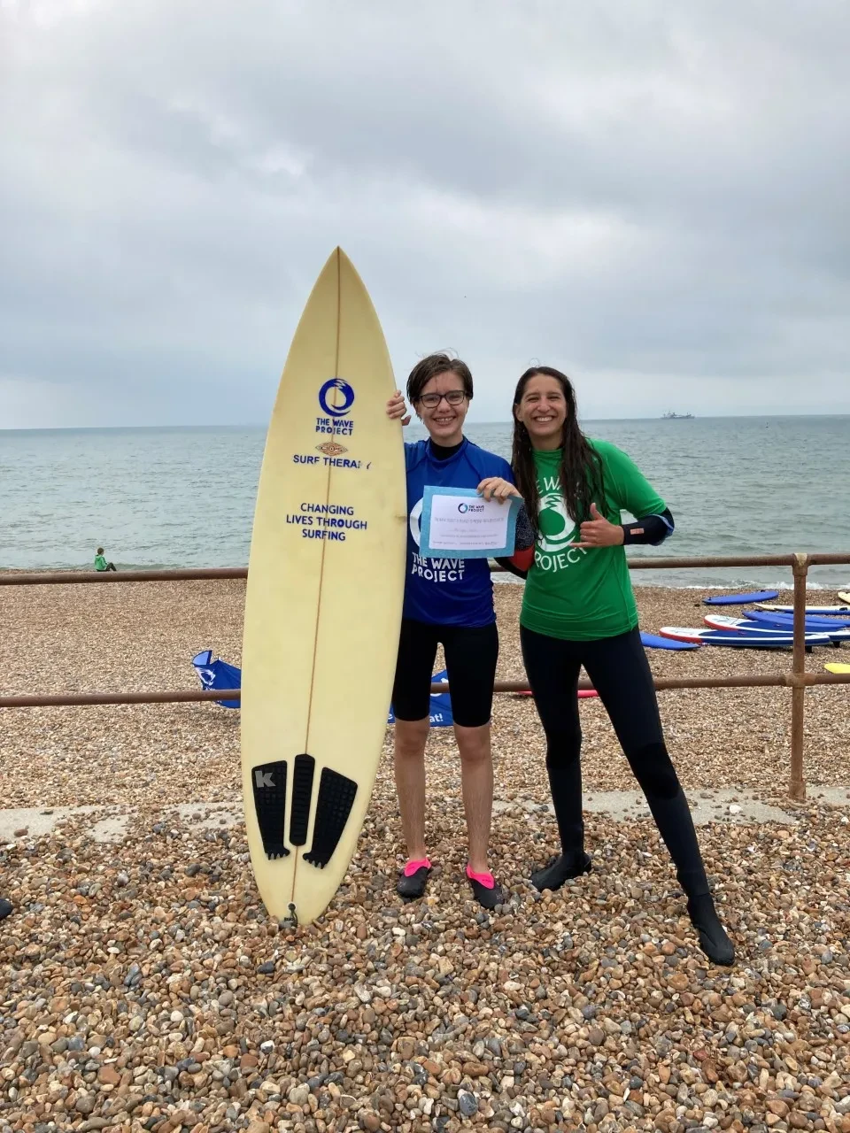 2 women on a pebble beach with sea and sky in the background. One is holding a surfboard and a certificate.
